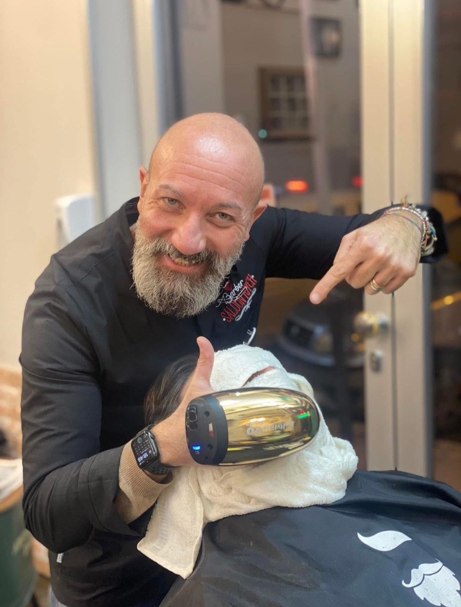 Barbiere calvo con barba grigia e casacca nera Saloon Kitch indica un cliente avvolto in un asciugamano bianco