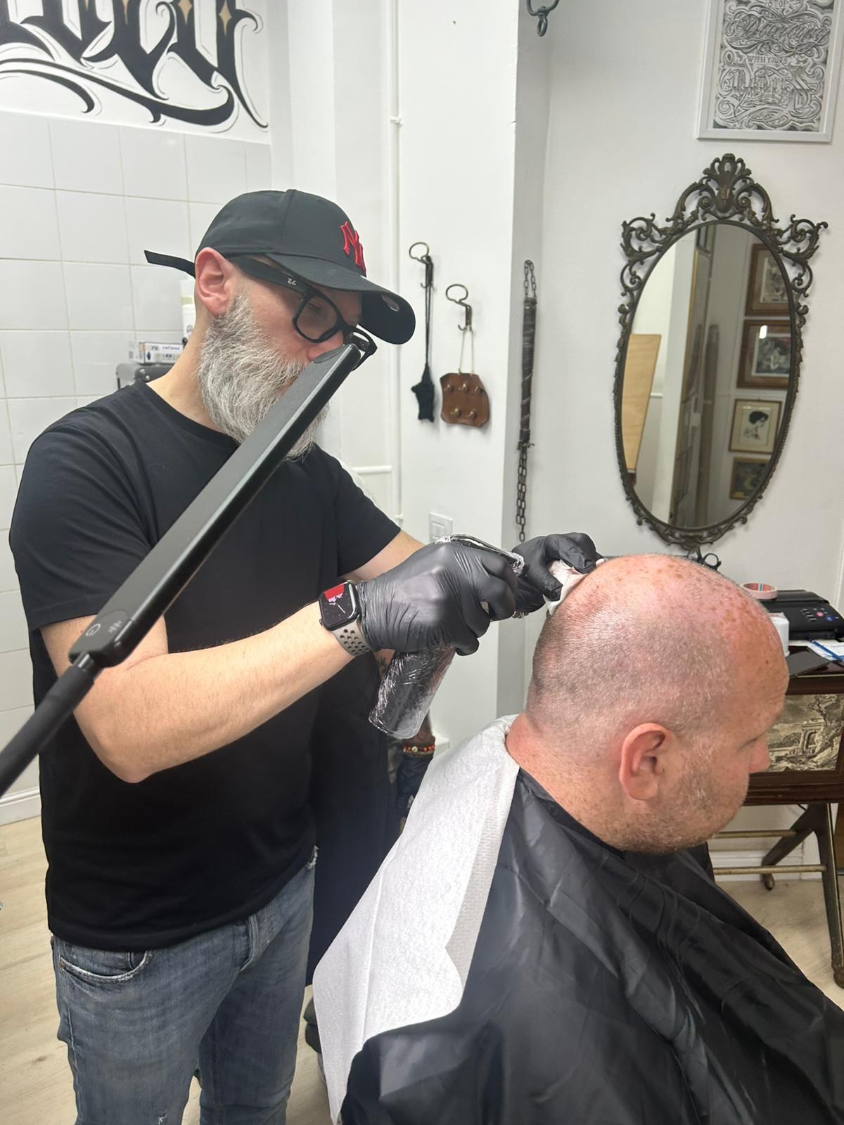 Operatore con barba grigia e guanti neri esegue tricopigmentazione su testa rasata di un cliente davanti a specchio ovale