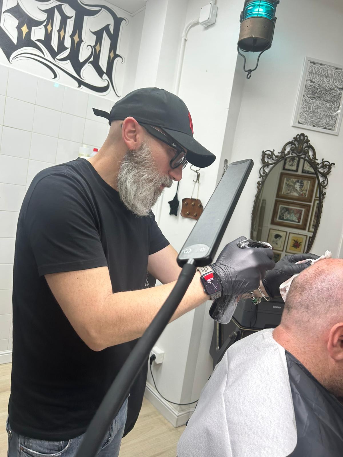 Operatore con barba grigia e guanti neri esegue tricopigmentazione sul capo di un cliente, accanto a uno specchio barocco